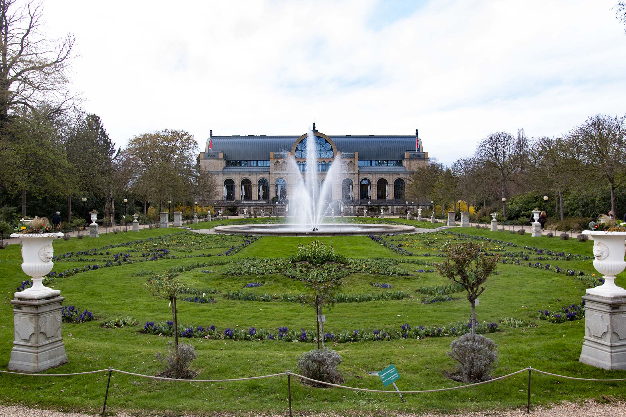 gepflegter Garten. Im Hintergrund eine Wasserfontäne und ein großes Haus.